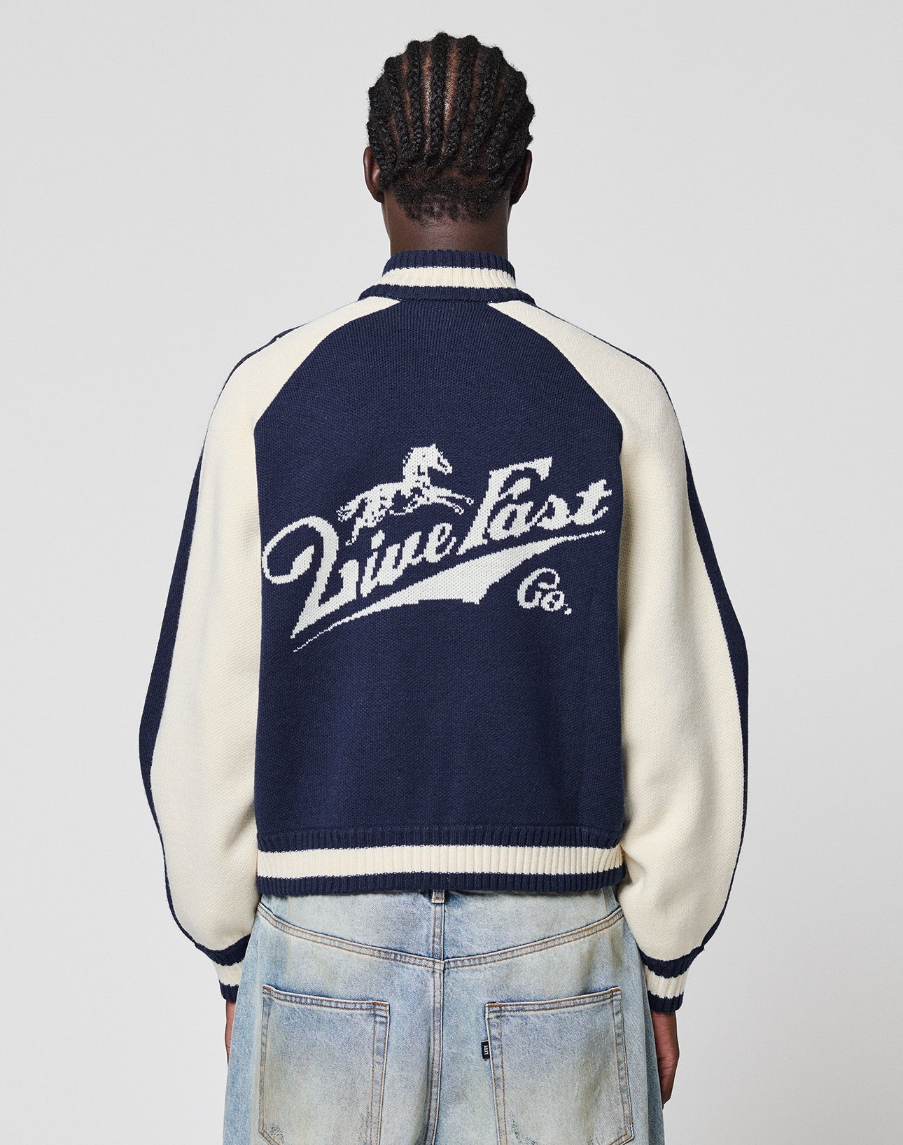 A person with braided hair, seen from behind, wears the Boxy Knit Zip Jacket by LIVE FAST DIE YOUNG with light blue oversize-fit jeans. The navy and cream jacket features a running horse logo and “Live Fast Co.” on the back. | model