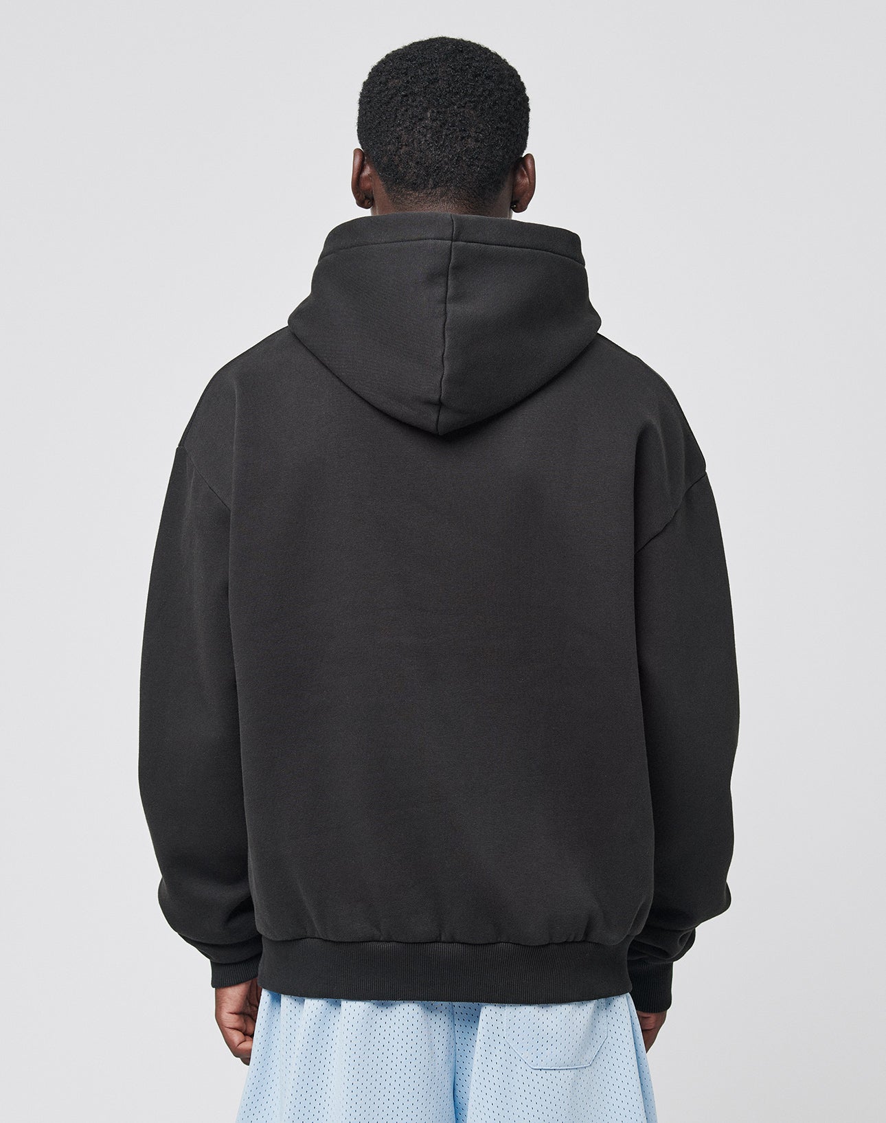 A person faces away from the camera wearing a LIVE FAST DIE YOUNG Live Fast College Hooded black oversize sweatshirt with the hood down and light blue mesh shorts, set against a plain gray background.