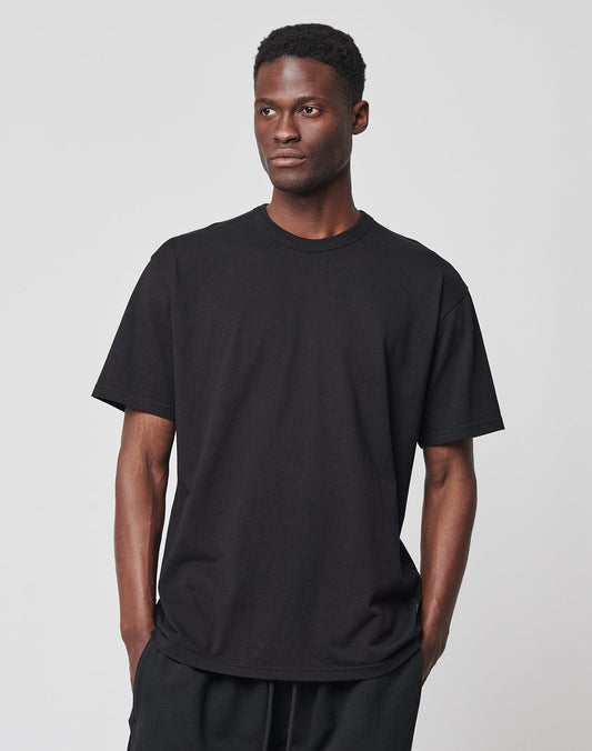 A man stands against a light gray background, wearing the LFDY Blank Tee by LIVE FAST DIE YOUNG— a Regular Fit black T-shirt made of 100% Baumwolle— and black pants, with his hands in his pockets and a neutral expression.