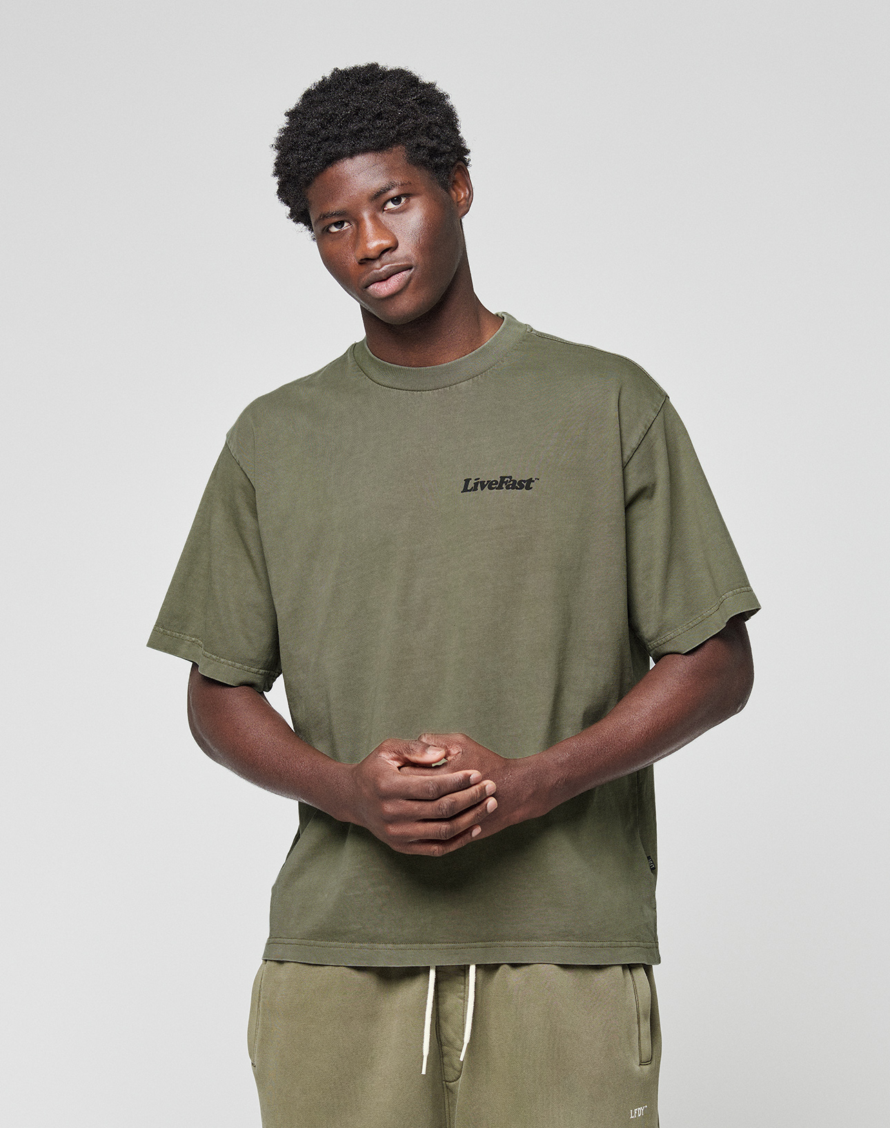 A young man faces the camera, hands loosely clasped, wearing a LIVE FAST DIE YOUNG Basic 210 Tee in olive green with front and back print. The oversize fit and 100% cotton offer comfort and style, paired with matching pants against a plain background.