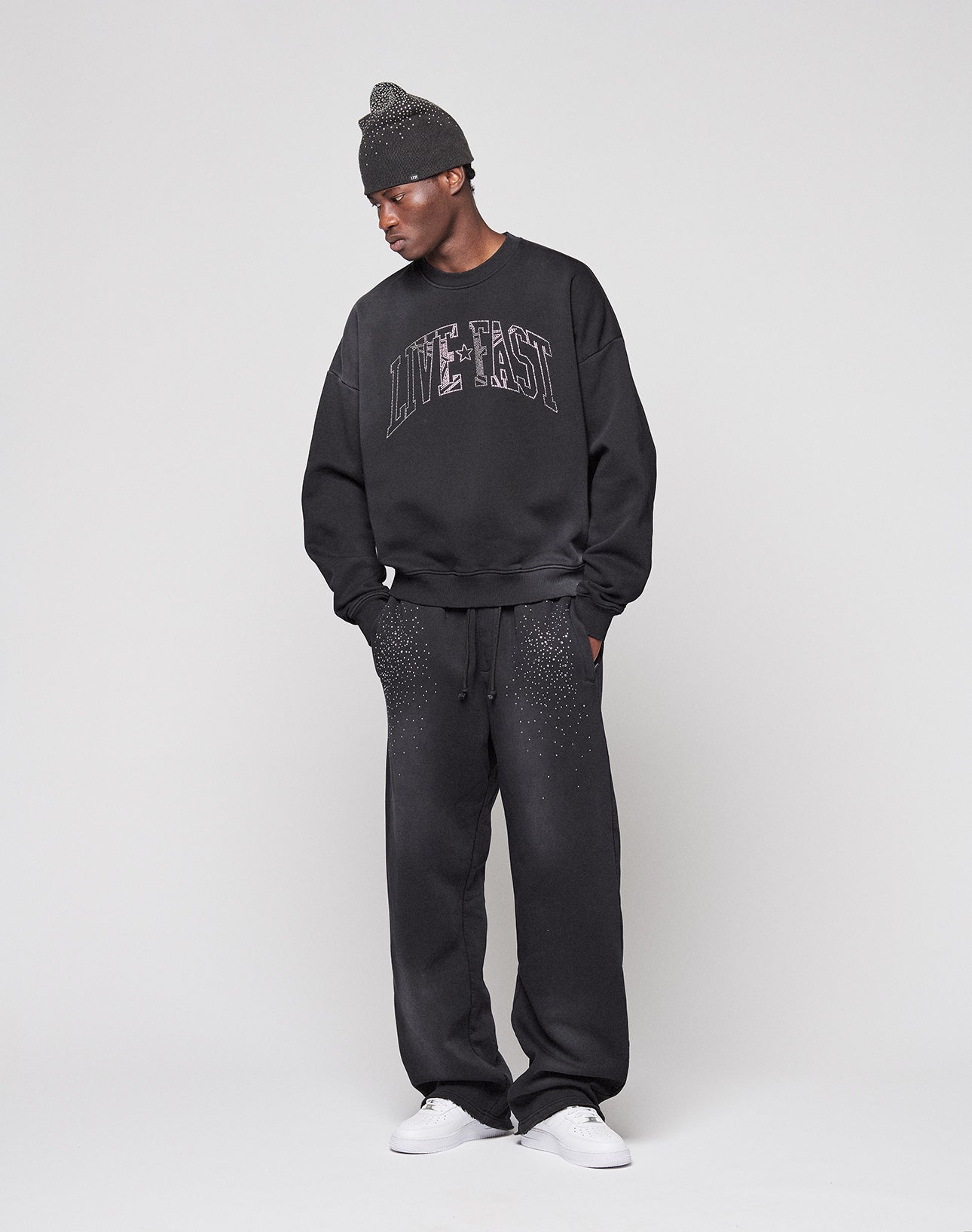 A person stands hands-in-pockets, wearing the LIVE FAST DIE YOUNG 'Live Fast Rhinestone Jogger'—a wide-fit, speckled black sweatpant—paired with a matching black beanie, sweatshirt featuring LIVE FAST text, and white sneakers.
