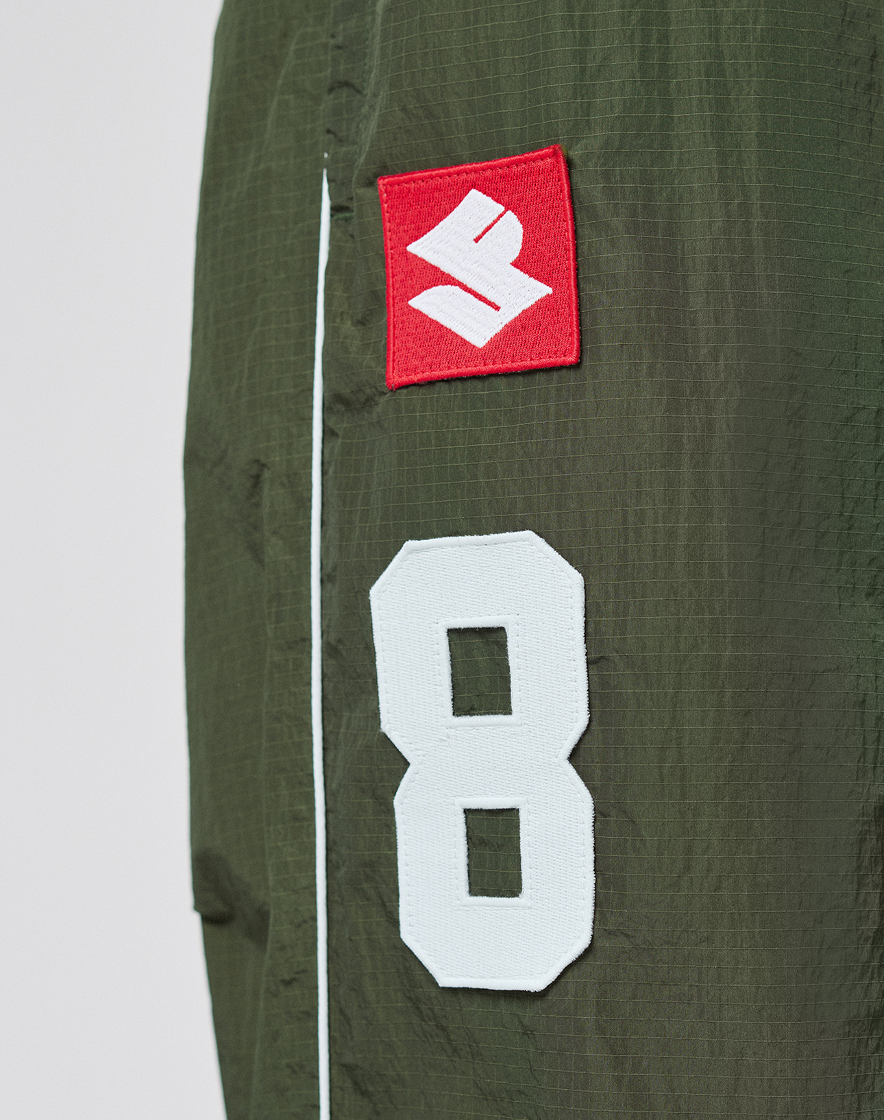 Close-up of olive green LFDY Nylon Trackpants by LIVE FAST DIE YOUNG, featuring a sewn white number 8 patch and red patch with a white S logo. A thin white stripe runs vertically beside the patches.