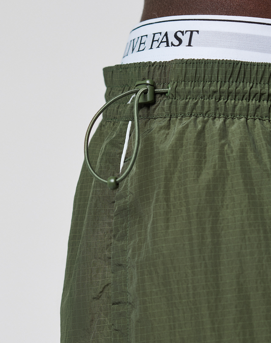 Close-up of LIVE FAST DIE YOUNG’s olive green LFDY Nylon Trackpants featuring a logo print, white waistband with “LIVE FAST” in black, wide fit, cinched elastic waist, polyamide blend fabric, and a drawstring with plastic toggle.