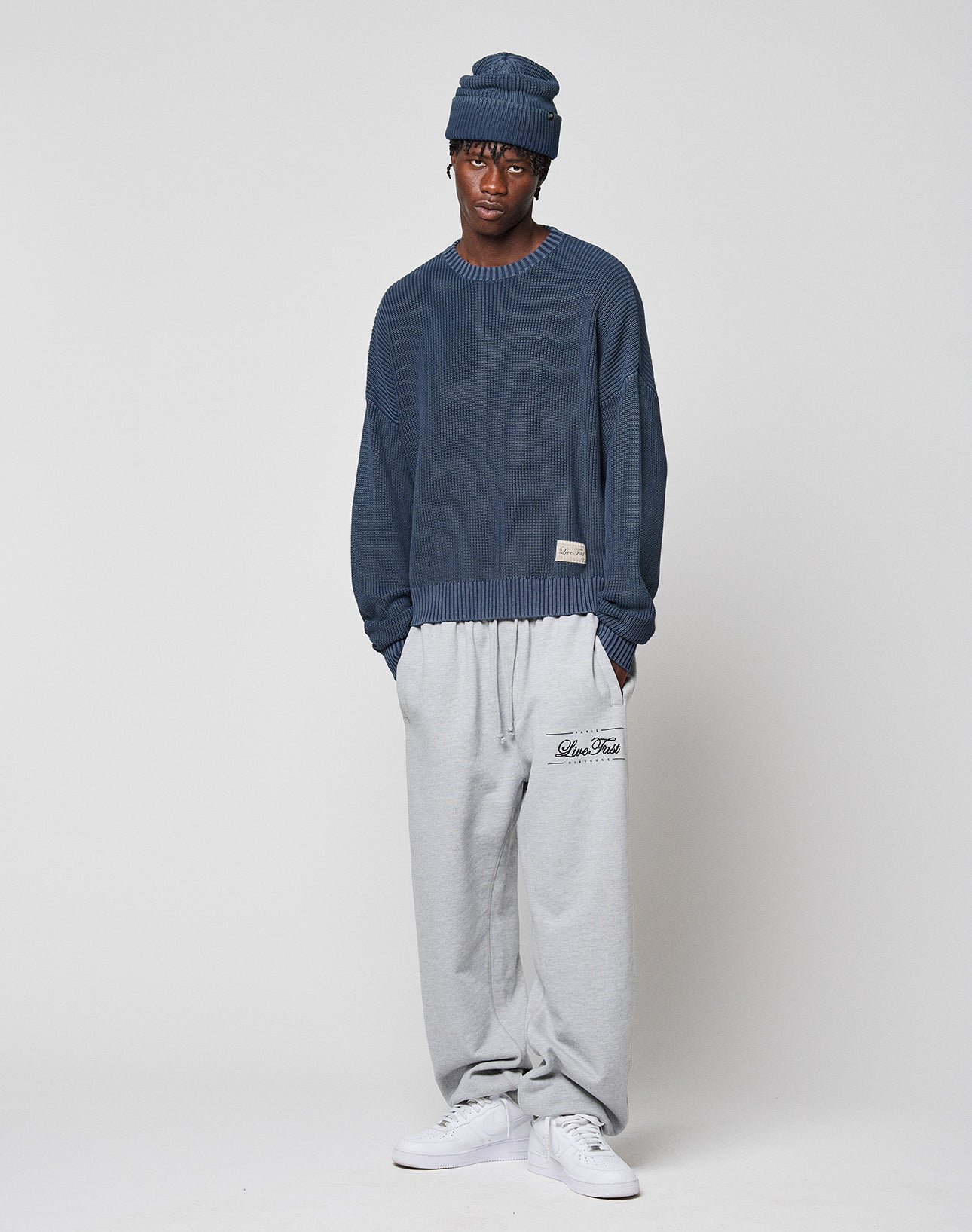 A person wears a LIVE FAST DIE YOUNG LFDY Knit Sweater, blue beanie, light gray sweatpants with left thigh text, and white sneakers while standing against a plain white background.