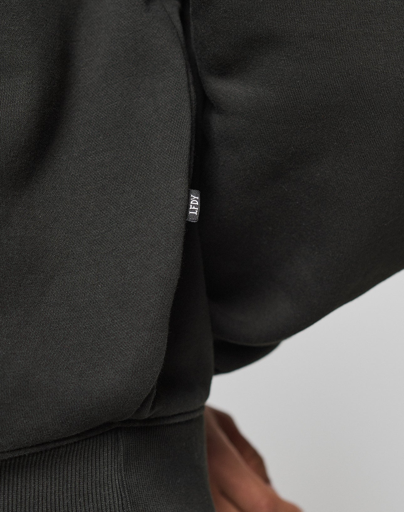 Close-up of a person wearing the Live Fast College Hooded by LIVE FAST DIE YOUNG in black, with a small rectangular tag on the side seam. Made of 65% Baumwolle and featuring an oversize fit. Part of their hand is visible at the bottom right.