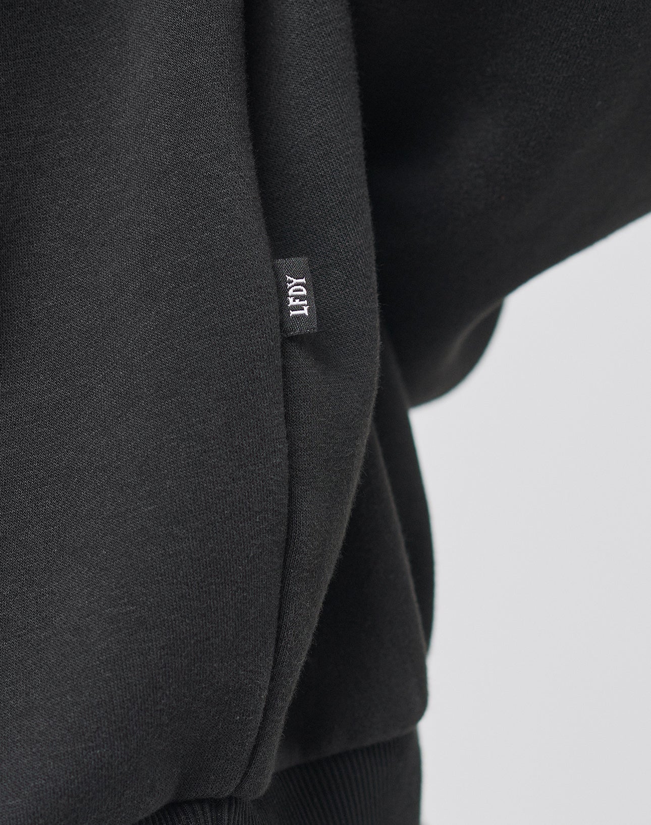 Close-up of a black Live Fast College Hooded by LIVE FAST DIE YOUNG, showing a small fabric tag with TIDY stitched in white. The soft material, likely cotton, features ribbed detailing at the bottom.