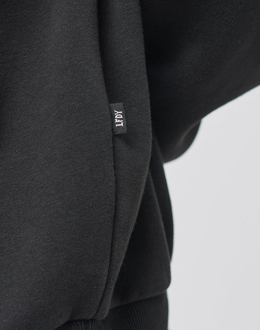 Close-up of a black Live Fast College Hooded by LIVE FAST DIE YOUNG, showing a small fabric tag with TIDY stitched in white. The soft material, likely cotton, features ribbed detailing at the bottom.