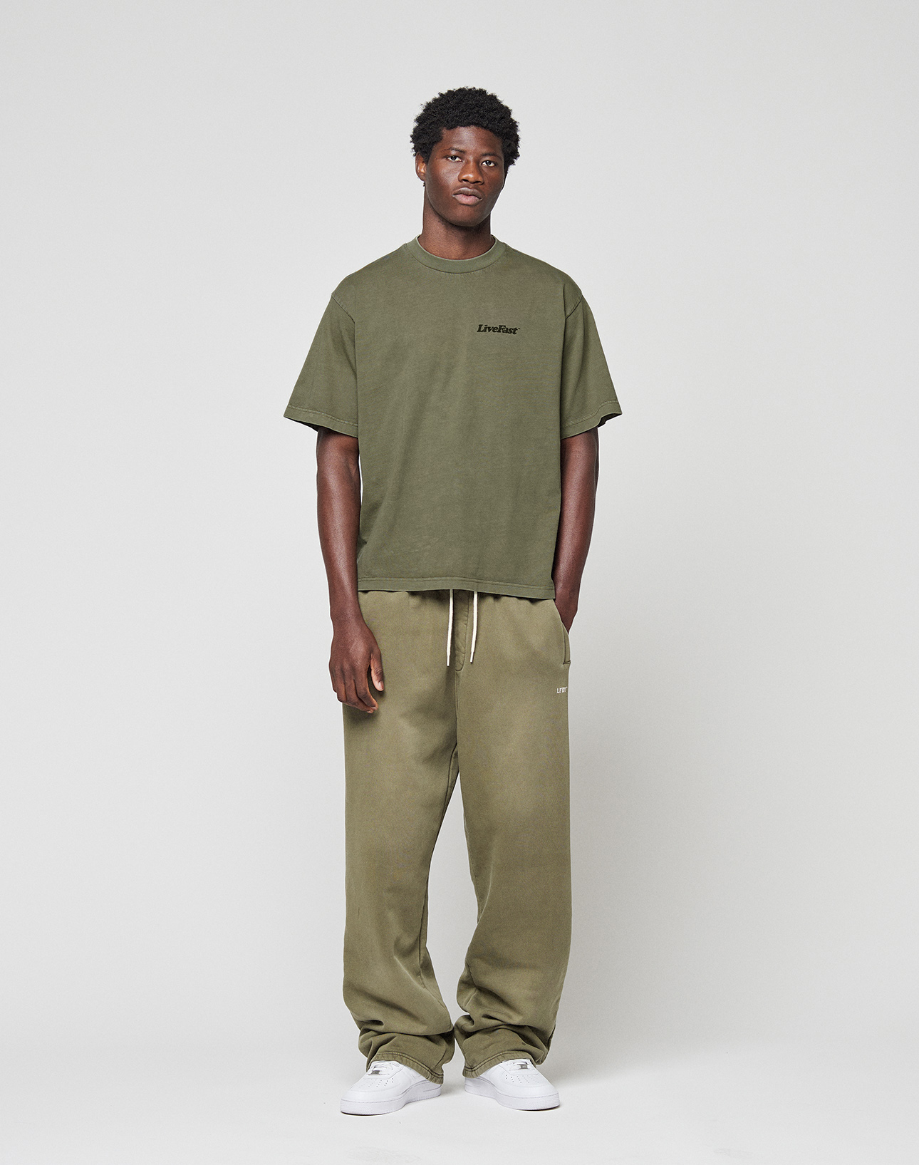 A man with short curly hair and a relaxed pose stands against a plain background in a LIVE FAST DIE YOUNG Basic 210 Tee, olive green and oversize fit, paired with matching loose sweatpants with drawstring and white sneakers.