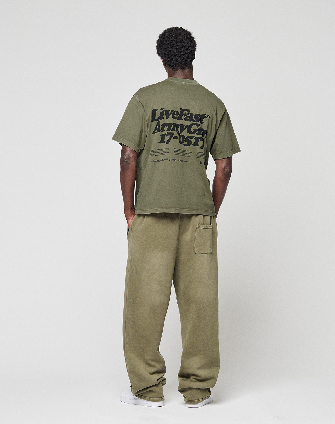 A person faces away, wearing a LIVE FAST DIE YOUNG Basic 210 Tee in olive green with black front and back print, paired with loose matching pants. The background is plain and light-colored.