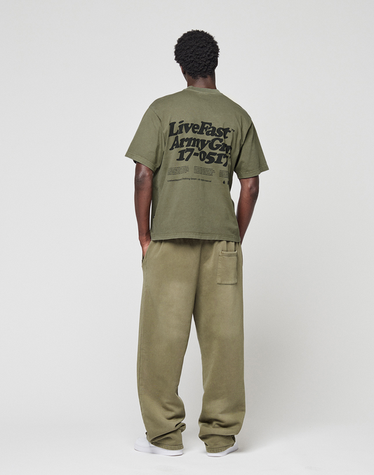 A person faces away, wearing a LIVE FAST DIE YOUNG Basic 210 Tee in olive green with black front and back print, paired with loose matching pants. The background is plain and light-colored.