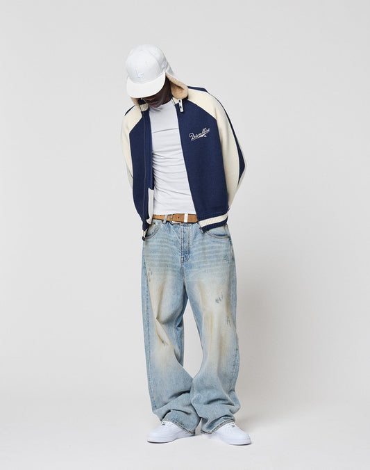 Wearing a Boxy Knit Zip Jacket by LIVE FAST DIE YOUNG in navy and cream, raglan sleeves, paired with a white cap, tee, baggy light blue jeans, and white sneakers, a person stands against a plain white background looking down with hands in pockets.