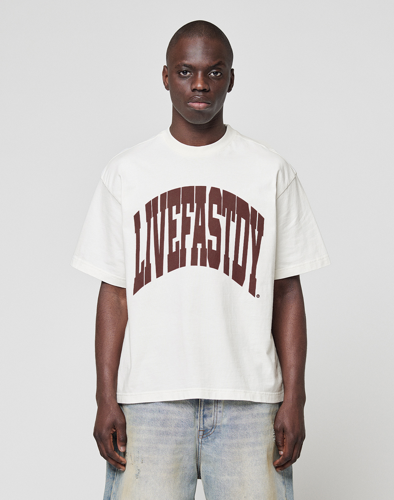 A person wears the LFDY Block Tee by LIVE FAST DIE YOUNG, an oversize fit white T-shirt with a bold dark red LIVEFASTDIE front print, paired with faded blue jeans. Made from 100% Baumwolle for premium comfort. | model