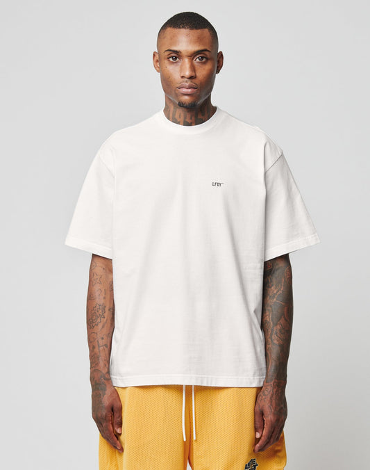 A person faces forward wearing the LIVE FAST DIE YOUNG Everybody Eats Tee in plain white 100% Baumwolle, featuring LIFT on the chest and yellow basketball shorts. Their tattooed arms and neutral expression stand out against a light gray background.