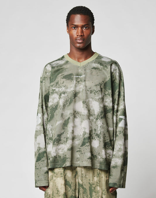 A man faces forward against a plain light background, wearing the LIVE FAST DIE YOUNG Camo Mesh Jersey—an oversized, long-sleeved green and beige camo shirt with a reflective print, number 23 on the sleeves—and light pants. | model