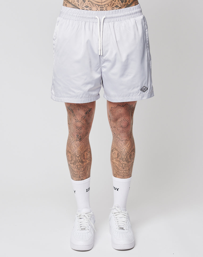A person wearing LIVE FAST DIE YOUNG Basic Satin Shorts with a logo patch, white socks, and white sneakers stands facing forward. Their tattooed legs are visible against a plain white background.