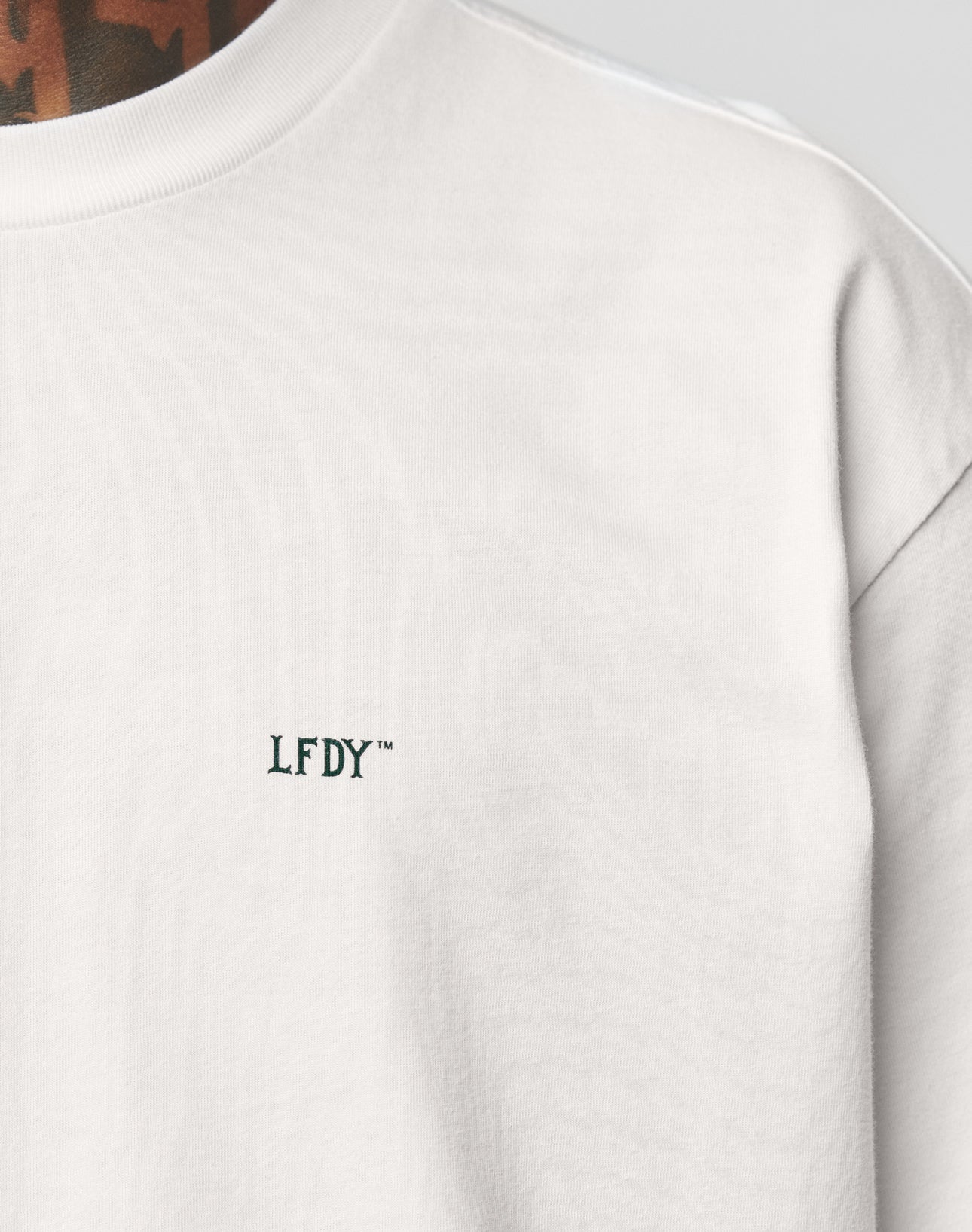 Close-up of a person in the LIVE FAST DIE YOUNG Everybody Eats Tee, an oversize white T-shirt with small, dark green LFDY embroidery on the left chest. Tattooed neck partly visible. Made from 100% Baumwolle (cotton).