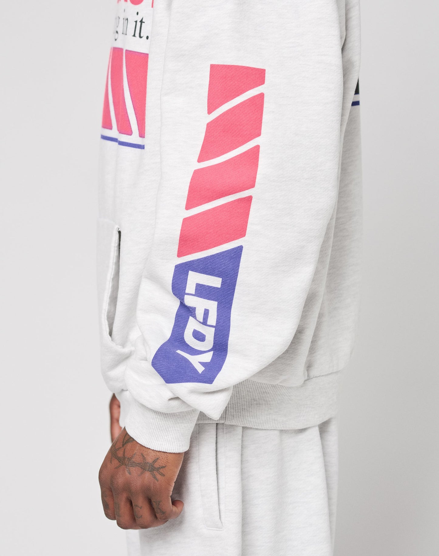 A person wears the LFDY Racing Hooded by LIVE FAST DIE YOUNG, an oversize fit light gray hoodie with red diagonal stripes and glossy LFDY on the sleeve. Their tattooed hand and matching gray Baumwolle Polyester pants are visible.