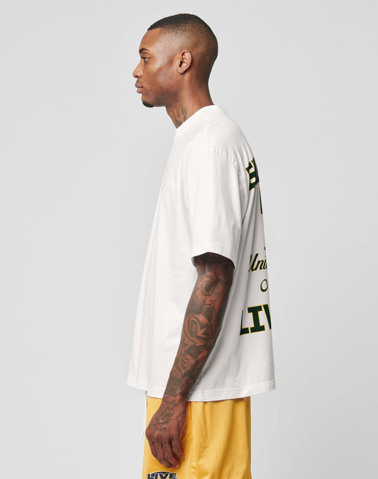 A man with a close-cropped haircut and tattoos on his left arm stands in profile wearing the LIVE FAST DIE YOUNG Everybody Eats Tee, an oversize fit white T-shirt with green and yellow backprint text, and yellow 100% Baumwolle athletic shorts.