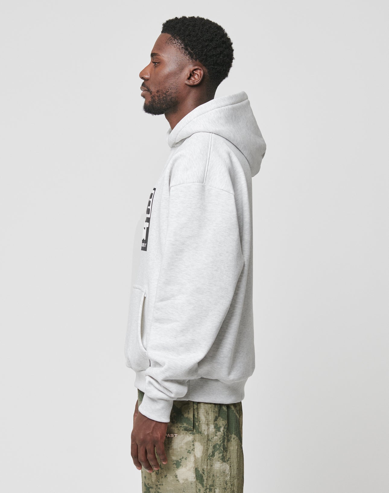 A man in profile wears the LIVE FAST DIE YOUNG Live Fast Box Hooded hoodie in light gray with an oversized fit and green camo pants made from soft Baumwolle, set against a plain light background.