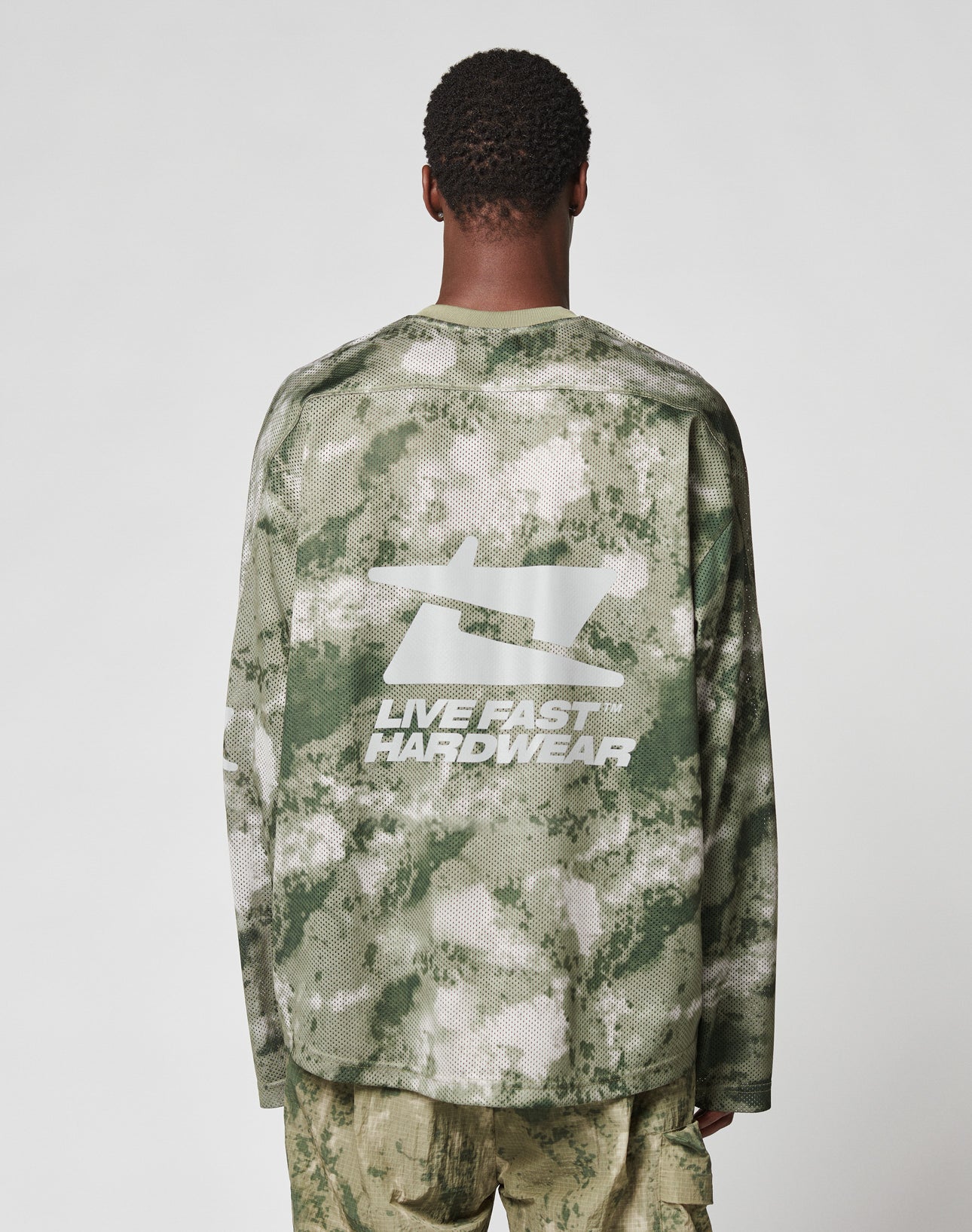 Someone wearing the LIVE FAST DIE YOUNG Camo Mesh Jersey, an oversize fit green and beige camouflage long-sleeve shirt with a large reflective print and LIVE FAST HARDWEAR text on the back, faces away from the camera against a plain background.