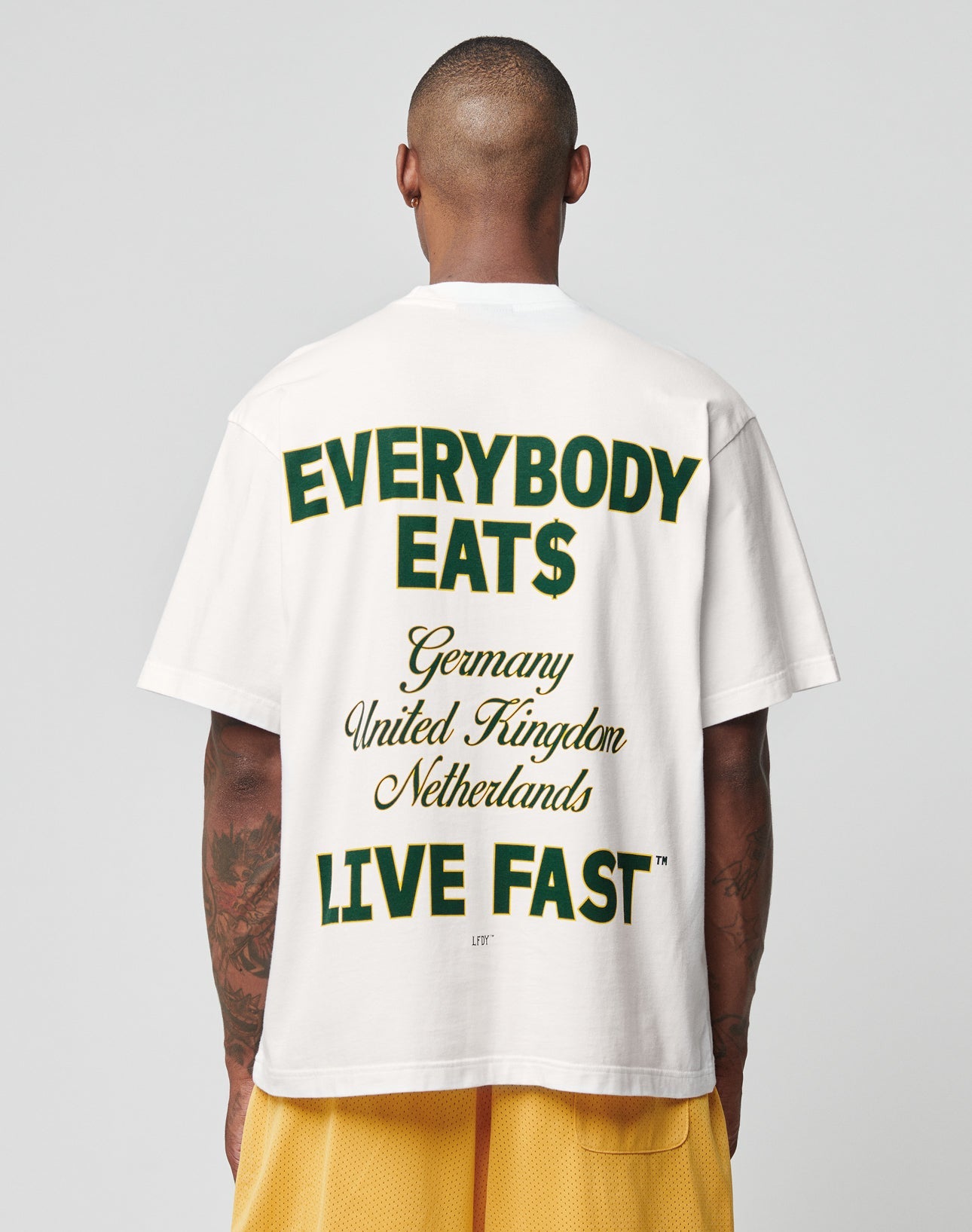 A person wears the LIVE FAST DIE YOUNG Everybody Eats Tee—an oversize fit white T-shirt made of 100% Baumwolle, featuring a green and yellow “EVERYBODY EAT$ Germany United Kingdom Netherlands LIVE FAST™” backprint—paired with yellow shorts.