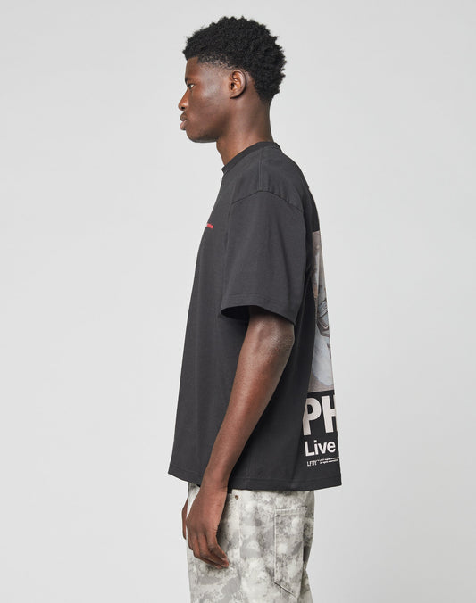 A young man in profile stands against a plain background wearing the black LFDY F*ck Phones Tee by LIVE FAST DIE YOUNG with light gray tie-dye pants. His arms hang relaxed and he looks ahead. Care instructions are included.