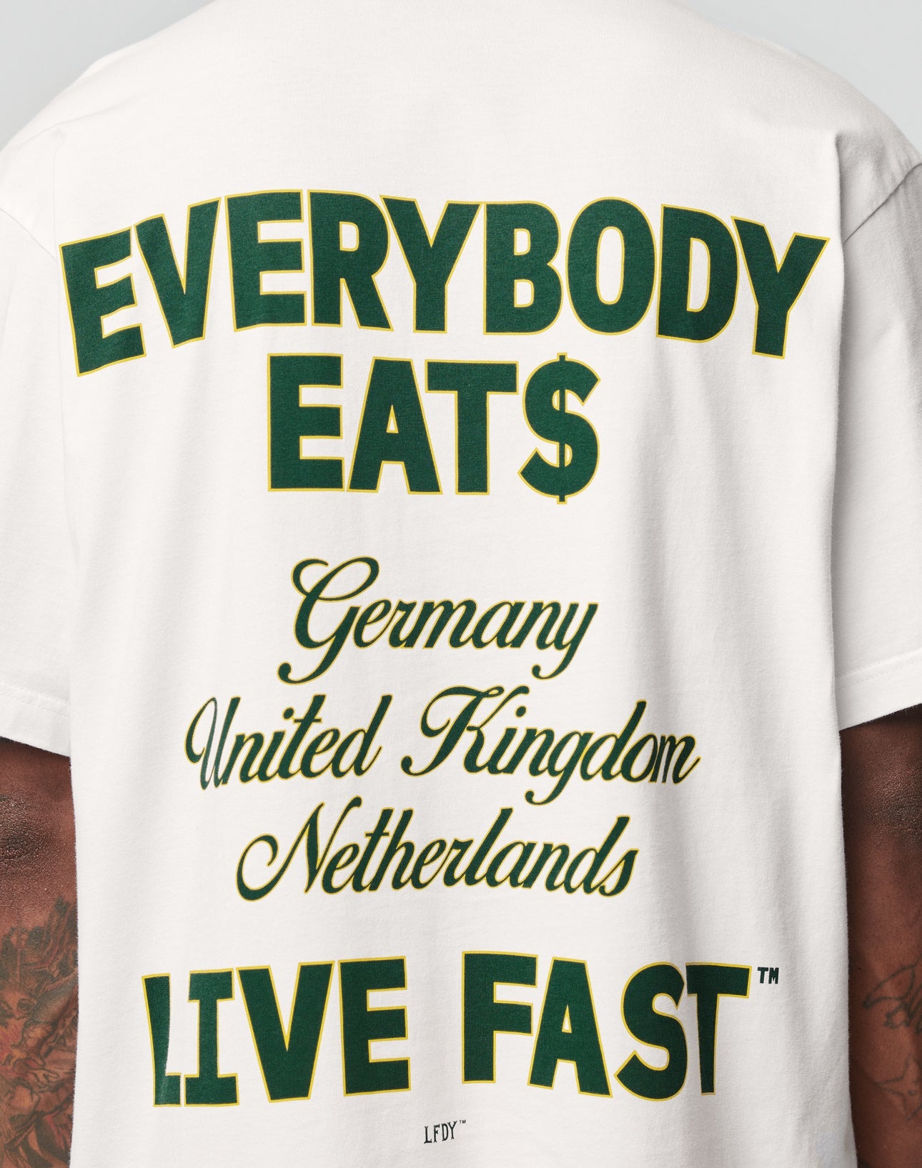 Someone wears the LIVE FAST DIE YOUNG Everybody Eats Tee, an oversized white 100% Baumwolle shirt with bold EVERYBODY EAT$ text, Germany/UK/Netherlands in the center, LIVE FAST at the bottom, plus a backprint.