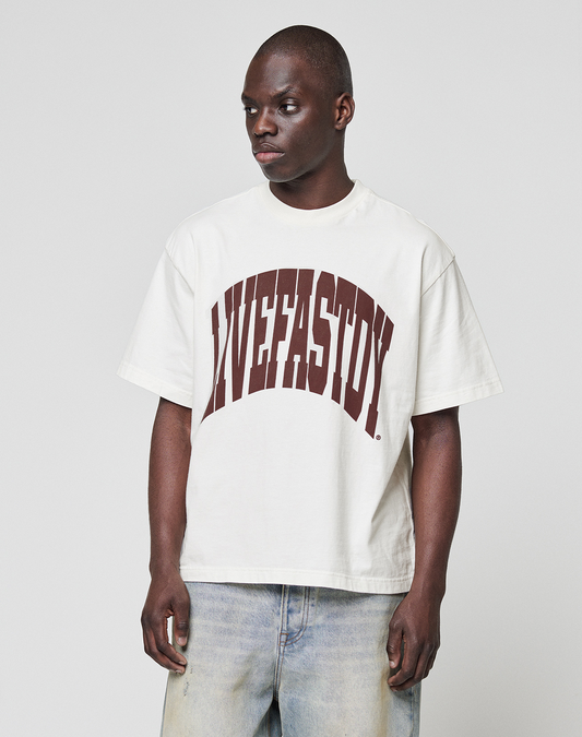 A person stands against a plain gray background wearing the LFDY Block Tee from LIVE FAST DIE YOUNG, a loose white T-shirt with a large brown "LIVEFASTDIE" front print, paired with light-washed jeans.