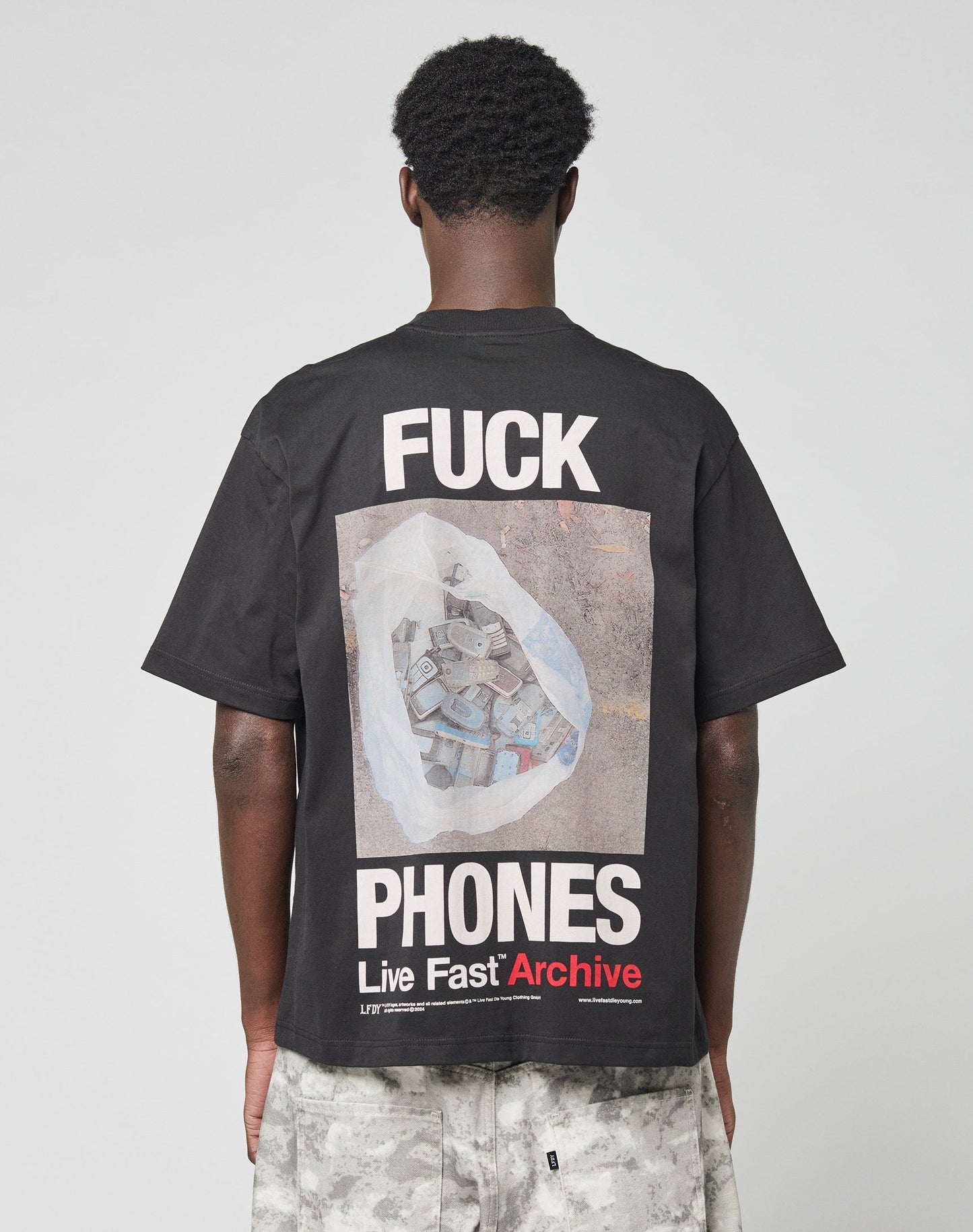A person is seen from behind wearing the LIVE FAST DIE YOUNG LFDY F*ck Phones Tee, an oversized black cotton shirt with FUCK PHONES and Live Fast Archive plus a bag of old phones graphic on the back. Care instructions are on the label.