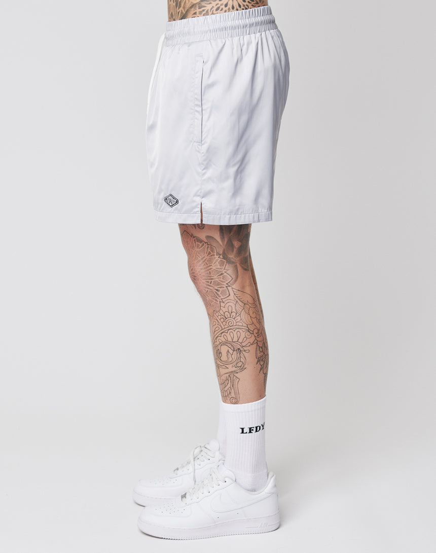 A person with leg tattoos is seen from the side wearing LIVE FAST DIE YOUNG Basic Satin Shorts, white socks with LEDY text, and white Nike sneakers, standing against a plain white background.