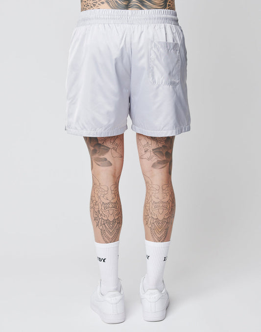 A person stands facing away, wearing LIVE FAST DIE YOUNG Basic Satin Shorts—shiny white with a logo patch and back pocket—plus white crew socks and sneakers, revealing detailed black tattoos on their legs against a plain white background.