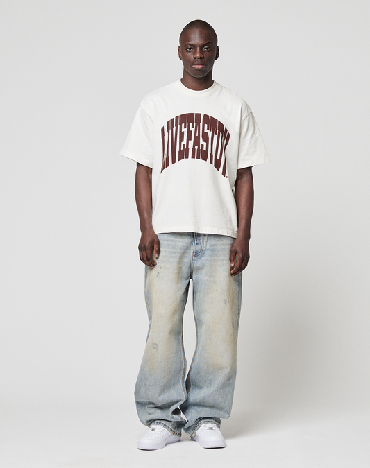 A person faces forward in a loose white LIVE FAST DIE YOUNG LFDY Block Tee with bold maroon "LIVESTOCK" frontprint, light blue baggy jeans, and white sneakers. The T-shirt has an oversize fit and is set against a plain white background.