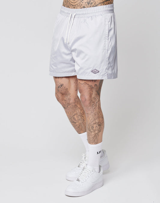 A person with tattooed legs wears LIVE FAST DIE YOUNG Basic Satin Shorts in light gray, featuring a subtle logo patch, paired with white crew socks and sneakers, standing against a plain white background.