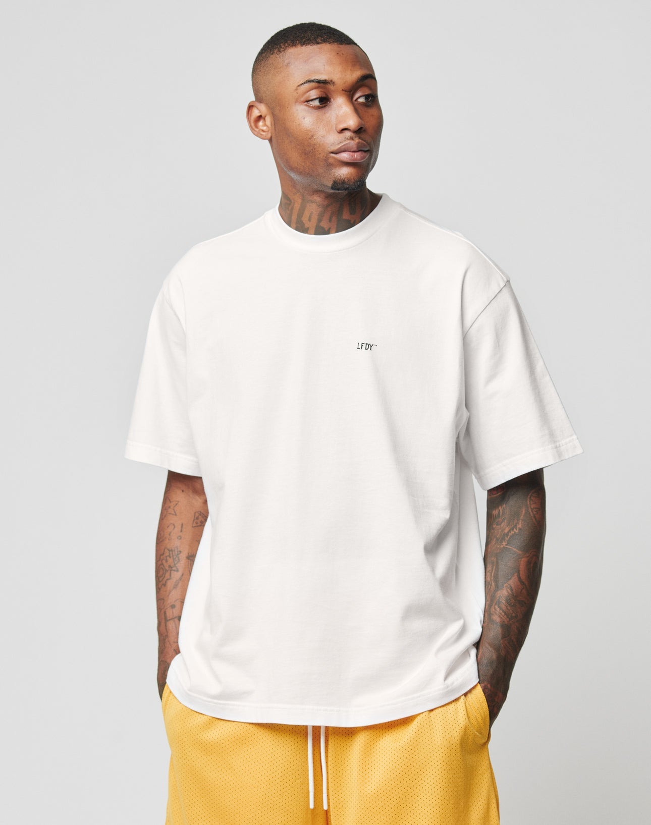 A man with short hair and visible tattoos wears the LIVE FAST DIE YOUNG Everybody Eats Tee in white and yellow athletic shorts, standing with his hands in his pockets against a plain light gray background.