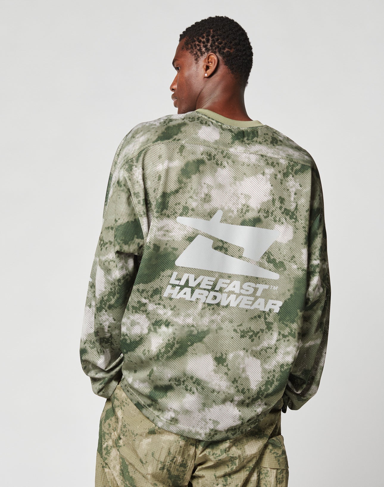 A person wearing a green and white Camo Mesh Jersey from LIVE FAST DIE YOUNG, featuring an oversized fit and a large reflective print on the back, stands against a plain light background.