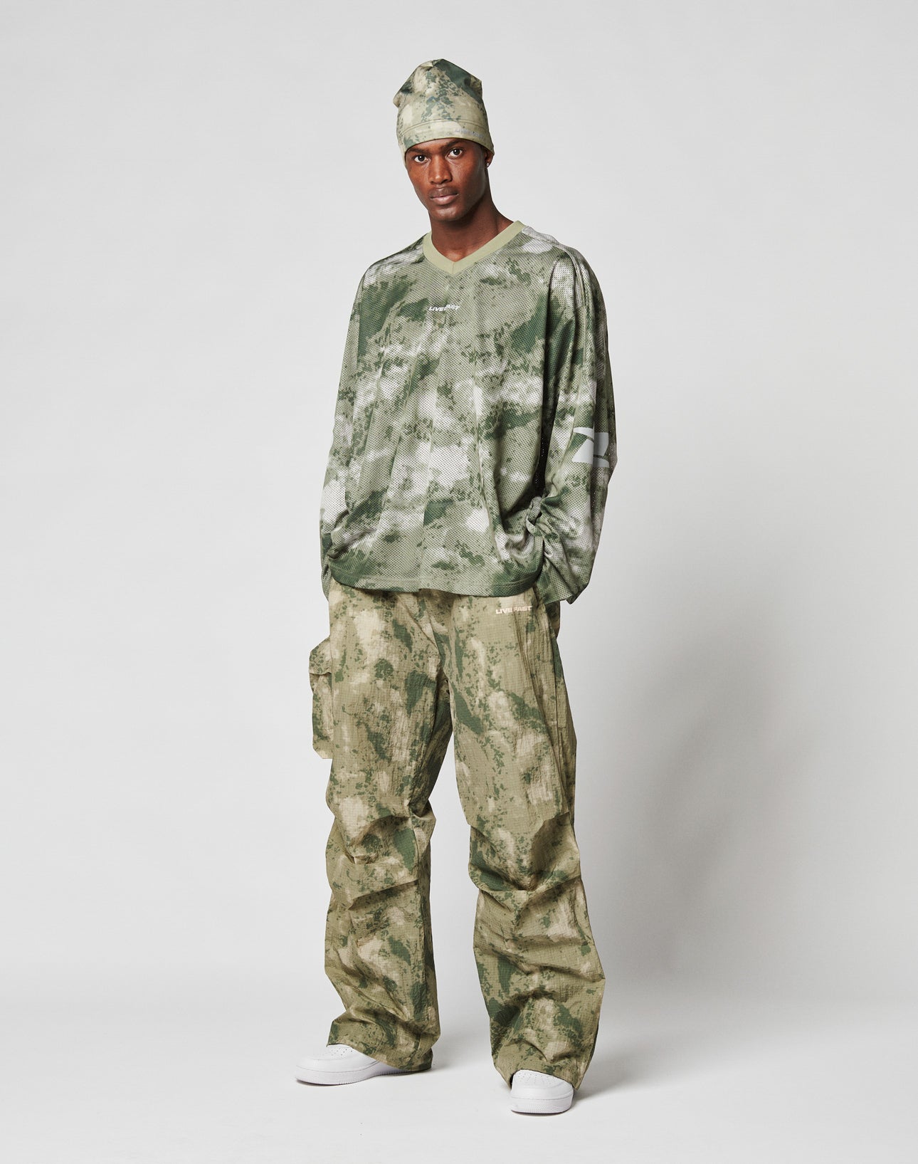 A man faces forward, wearing the LIVE FAST DIE YOUNG Camo Mesh Jersey—an oversize green and beige camo-patterned shirt and pants set made from 100% polyester—styled with a matching headscarf and white sneakers against a plain light background.
