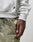 A close-up shows a person wearing the LIVE FAST DIE YOUNG Live Fast Box Hooded in light grey Baumwolle with an oversized fit, paired with camouflage pants. A small black label is visible on the sweatshirt as their hand rests by their side.
