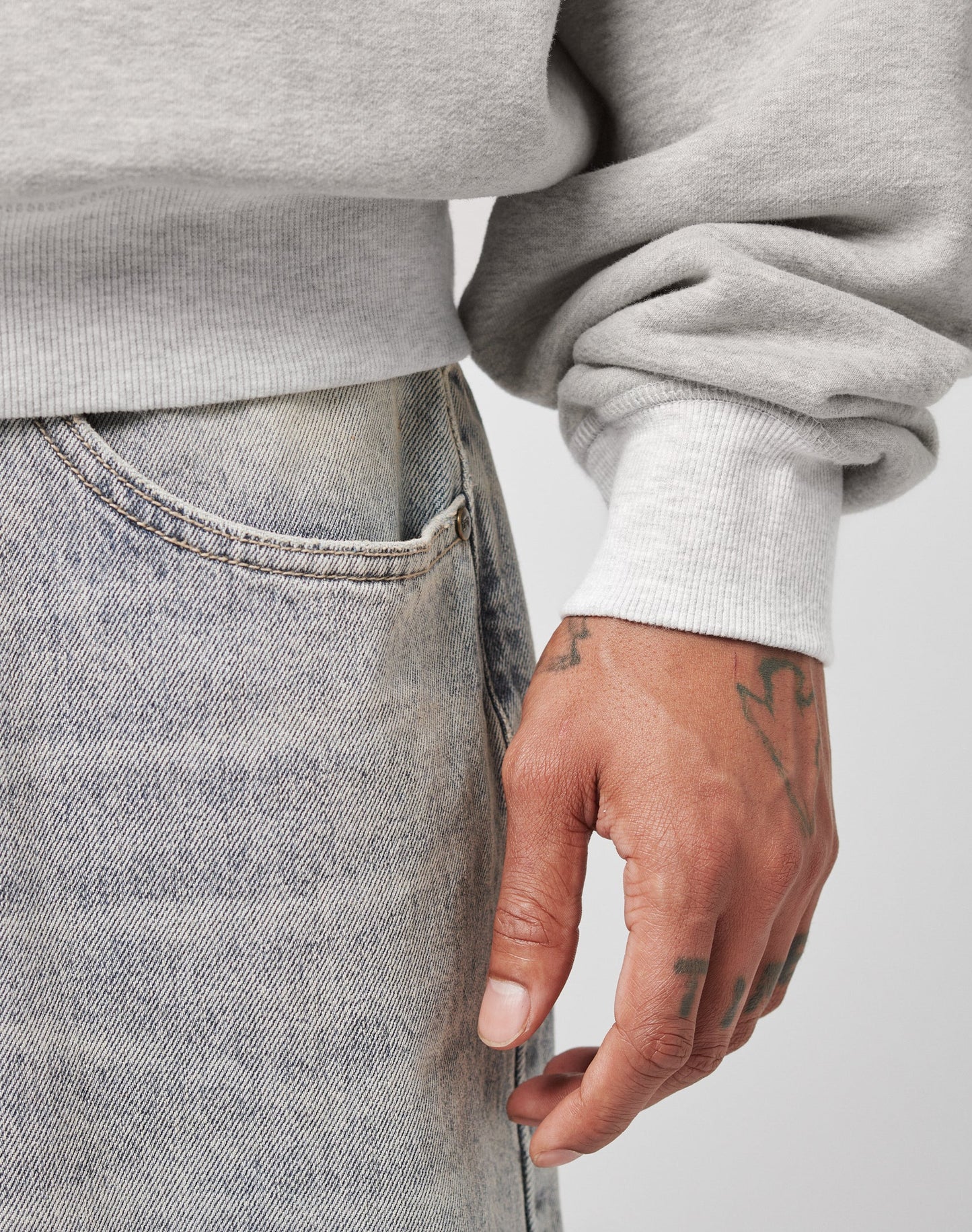 A close-up of a tattooed hand wearing the LIVE FAST DIE YOUNG LFDY Script Sweater in light gray with raglan sleeves and oversize fit, paired with faded blue jeans. The relaxed hand rests beside the jeans pocket.