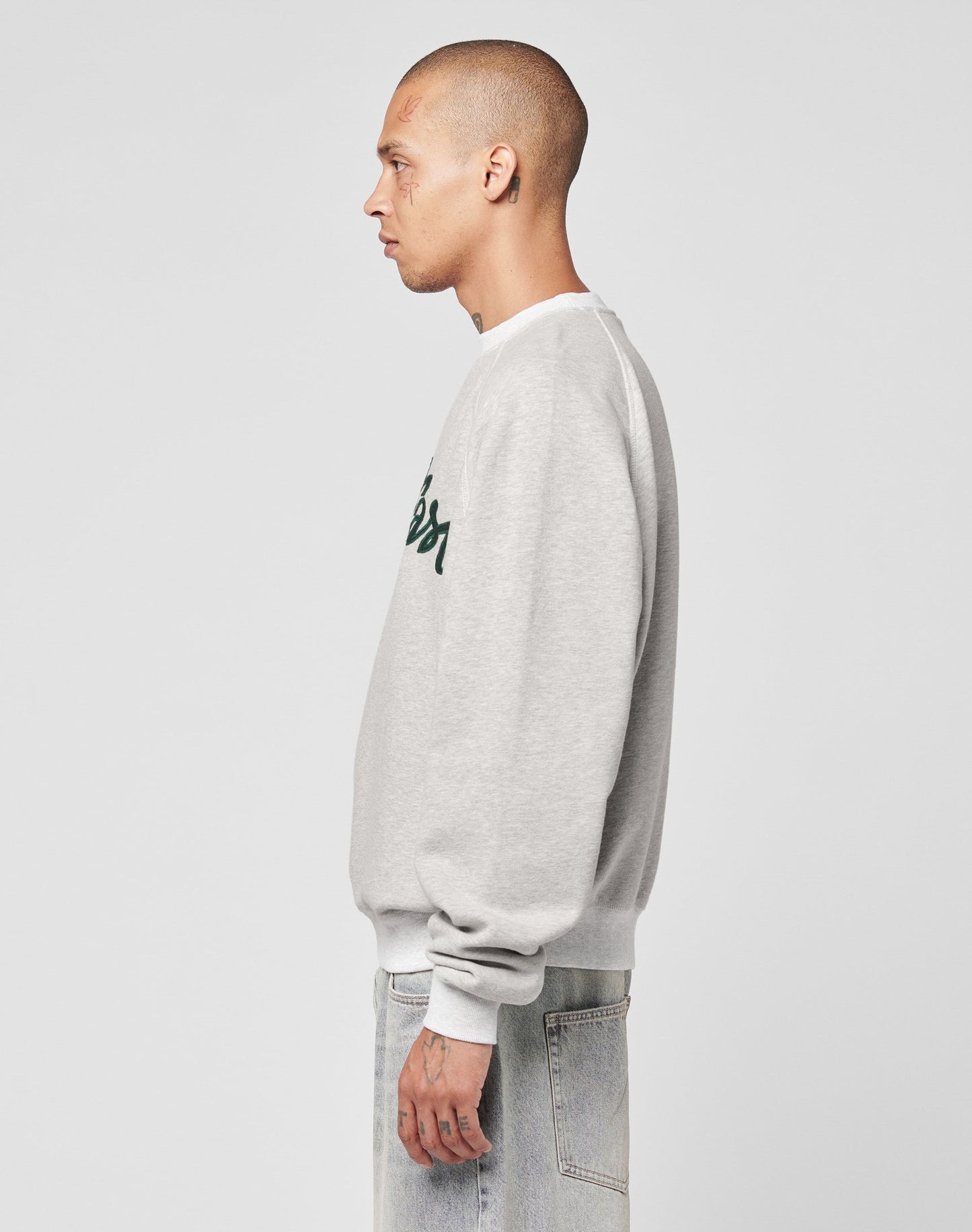 A person with a shaved head, wearing the LFDY Script Sweater by LIVE FAST DIE YOUNG in light gray and light blue jeans, stands in profile facing left against a plain white background.