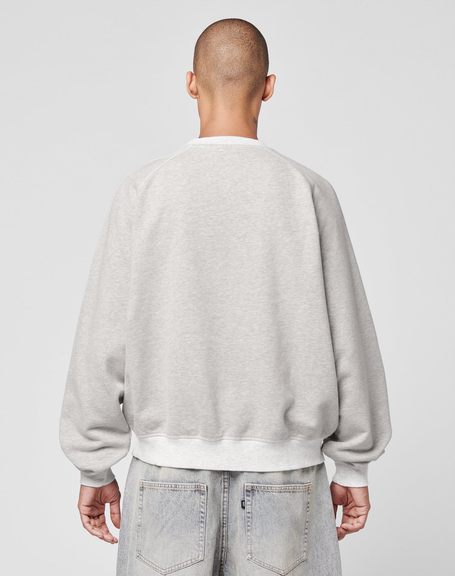 A person with a shaved head is shown from behind, wearing the LIVE FAST DIE YOUNG LFDY Script Sweater in light gray with raglan sleeves and light blue denim jeans against a plain, light background.