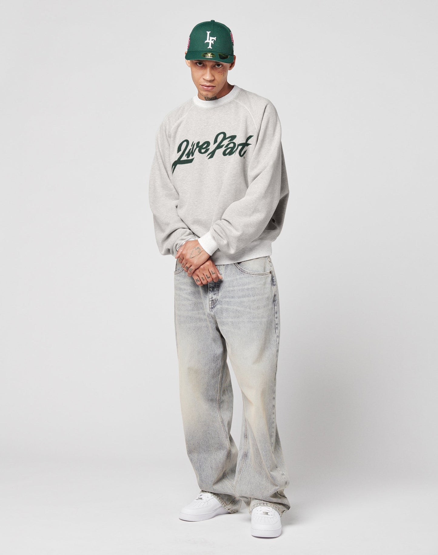 A person in an oversize fit, grey LFDY Script Sweater by LIVE FAST DIE YOUNG with raglan sleeves and green Love Pat script, loose faded jeans, a green baseball cap, and white sneakers stands against a plain white background.