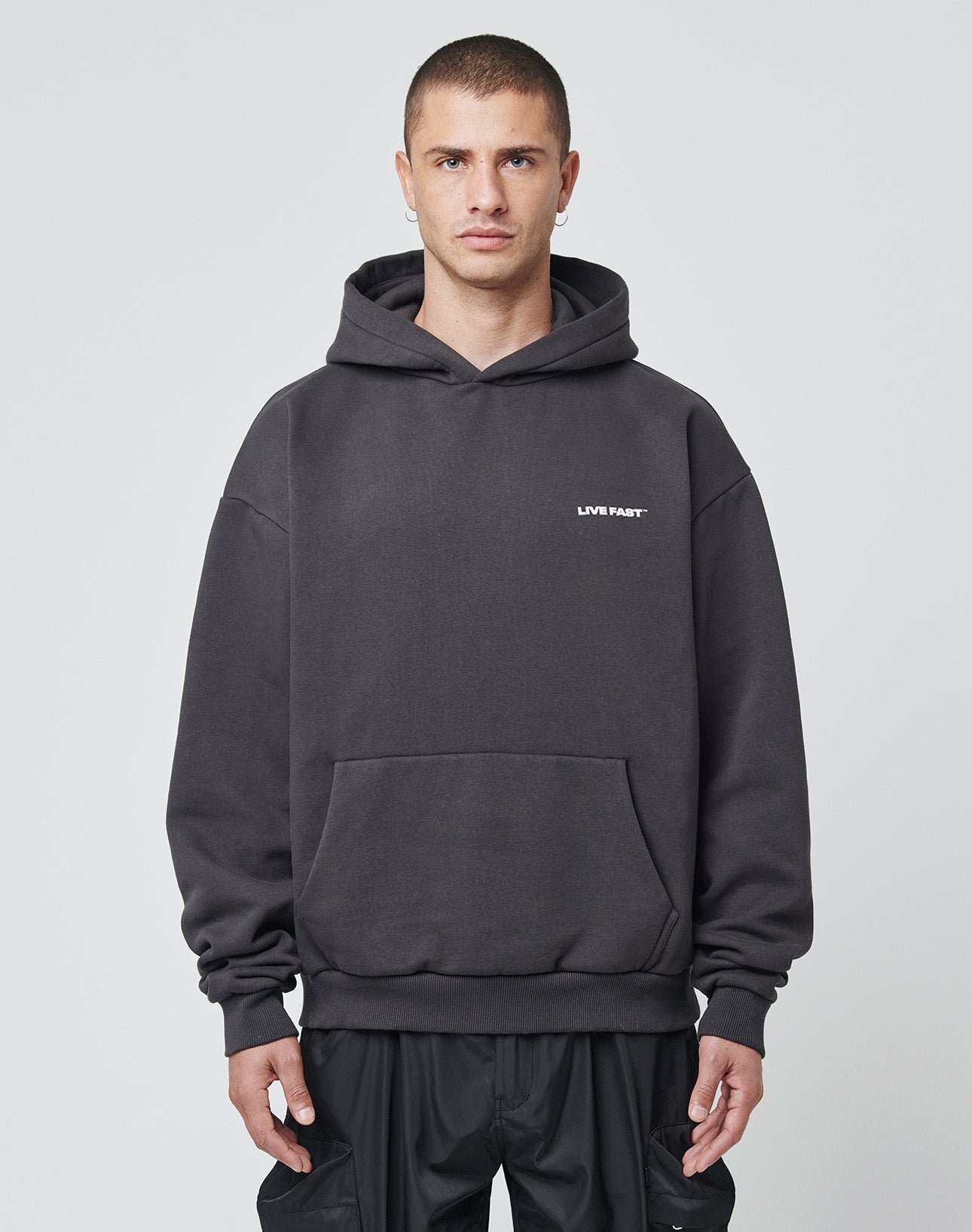 A person with short hair wears an oversized, dark gray LFDY Cinema Hooded by LIVE FAST DIE YOUNG, made from cotton-polyester and featuring small white LIVE FAST text on the left chest, standing before a light gray background.