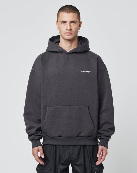 A person with short hair wears an oversized, dark gray LFDY Cinema Hooded by LIVE FAST DIE YOUNG, made from cotton-polyester and featuring small white LIVE FAST text on the left chest, standing before a light gray background.