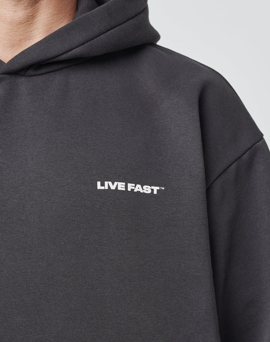 Close-up of a person wearing the LFDY Cinema Hooded by LIVE FAST DIE YOUNG, a black oversize fit hoodie with LIVE FAST™ in white on the chest. Only the upper torso and part of the hood are visible.