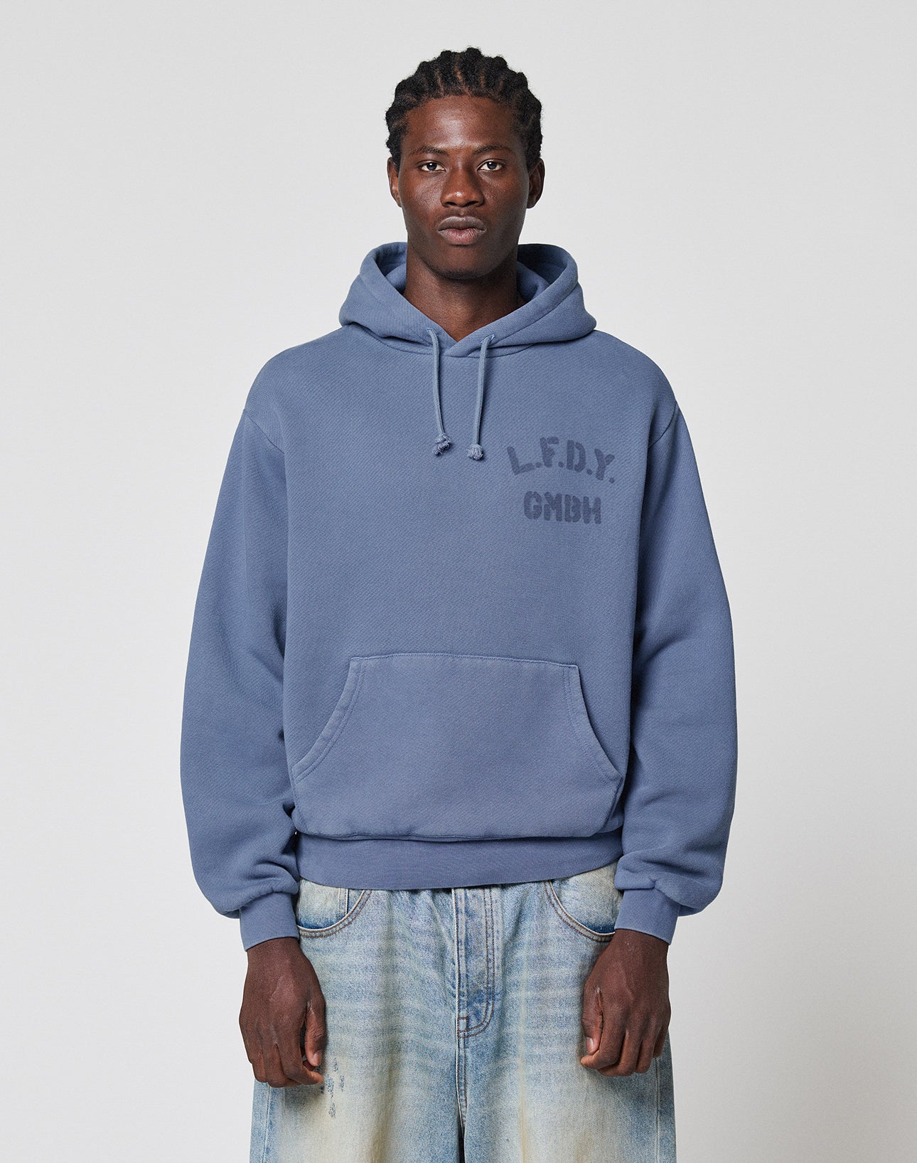 A man faces forward in a LIVE FAST DIE YOUNG LFDY GmbH Hooded with drawstring hood and chest print, paired with light blue faded jeans, against a plain gray background. | model