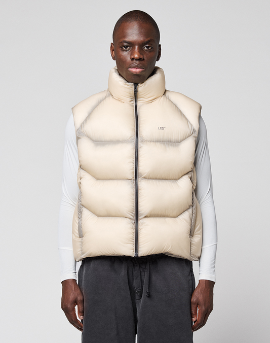 A man faces forward against a light gray background, wearing the LIVE FAST DIE YOUNG Live Fast Puffer Vest in light beige over a white long-sleeve shirt and dark drawstring pants. | model