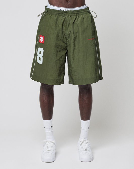 A person in LFDY Nylon Trackshorts by LIVE FAST DIE YOUNG—olive green, wide fit with a white number 8 and red logo-print—paired with white socks and sneakers, stands against a plain light background.