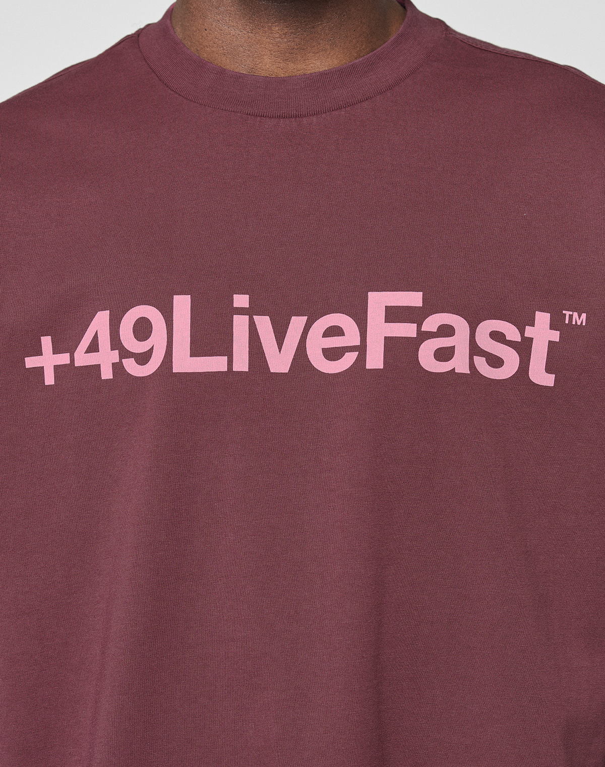 A person wearing the LIVE FAST DIE YOUNG Basic +49 Tee, an oversize fit maroon shirt made from 100% Baumwolle, featuring a pink +49LiveFast™ front print across the chest.