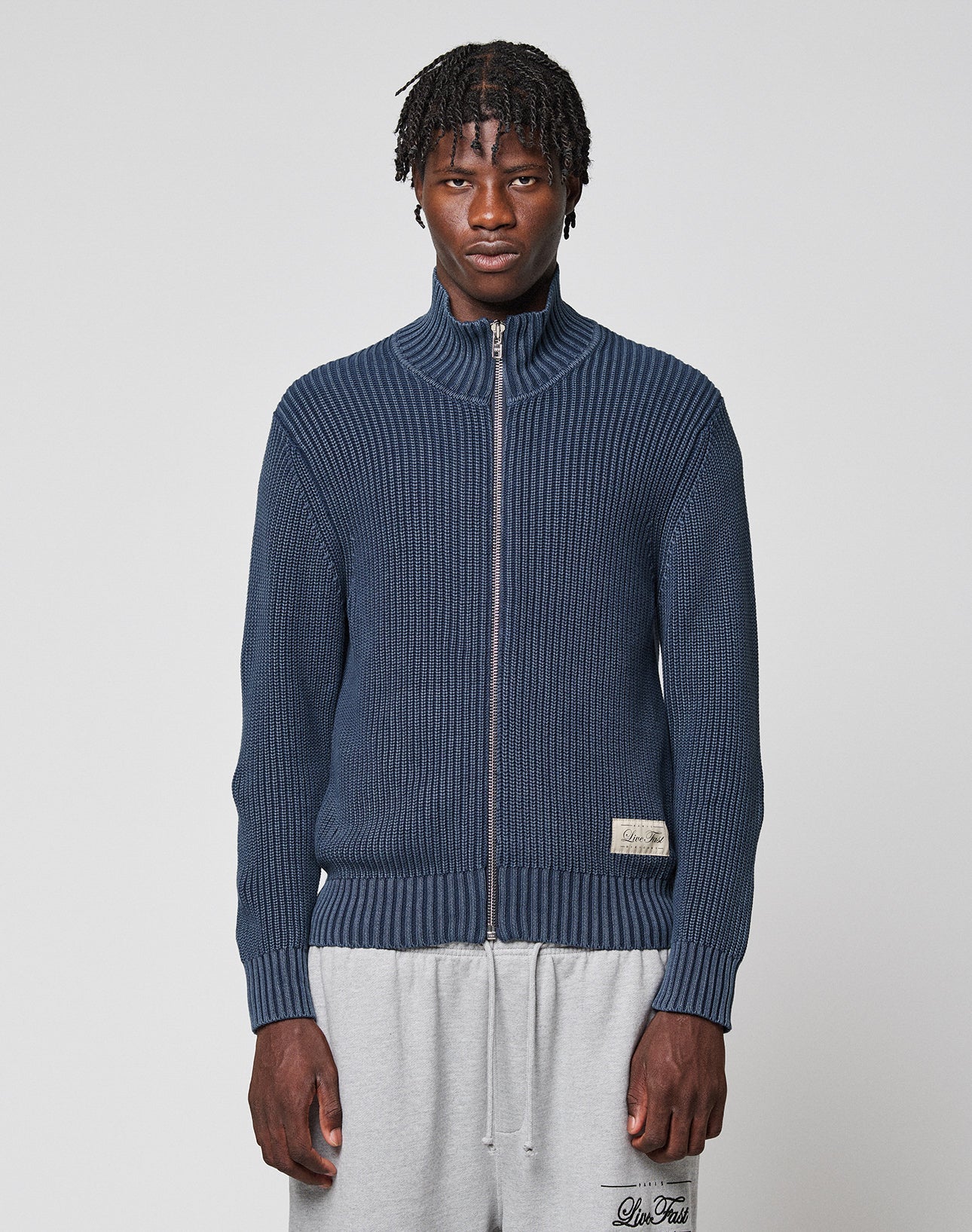 A man with short, twisted hair wears the LFDY Knit Jacket by LIVE FAST DIE YOUNG—a navy ribbed zip-up sweater with stand-up collar—and light gray sweatpants, standing against a plain light gray background.