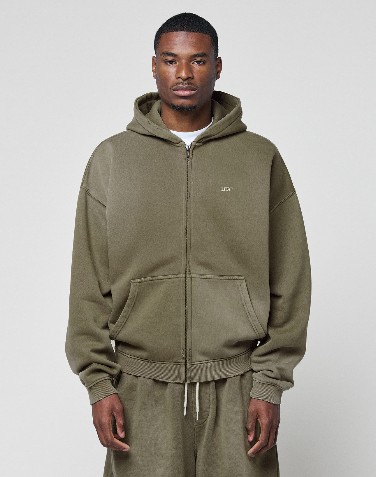 A man faces forward against a plain light background, wearing the LIVE FAST DIE YOUNG Basic Hooded Zip in olive green with a faded finish and matching sweatpants, his hands relaxed at his sides. | model